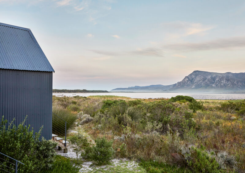 Modern gray building beside a coastal landscape with shrubs, mountains, and a serene sky in South Africa.