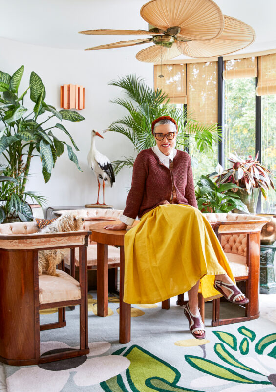 A woman in a yellow skirt and brown cardigan sits on a wooden table in a bright, plant-filled room with a stork statue.