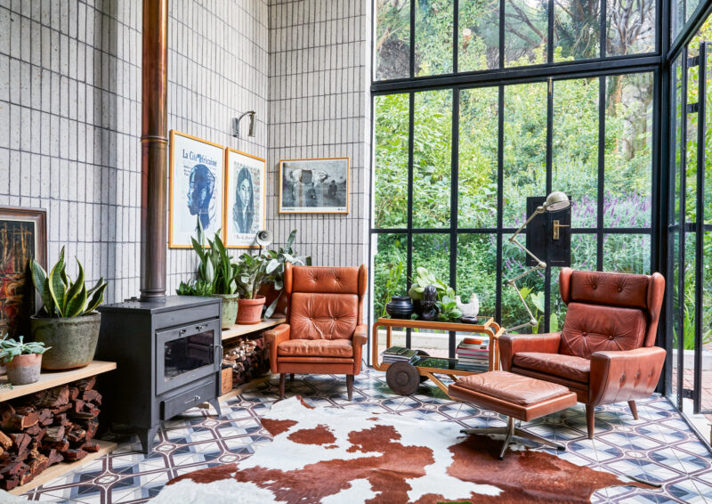 Cozy interior of Bureaux House in Cape Town, featuring leather armchairs, plants, and a wood stove against a glass wall.