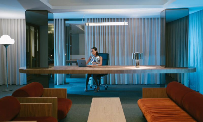 A woman in a stylish outfit sits at a modern desk with a laptop, surrounded by contemporary furniture and soft lighting.