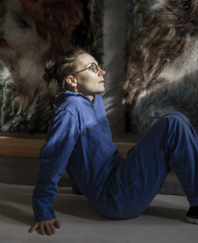 A woman in a blue jumpsuit sits against a wall, gazing upward, with textured artworks behind her.