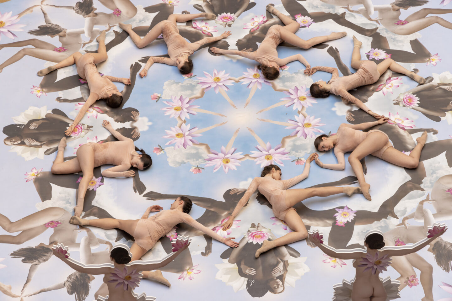 A circular arrangement of dancers in nude costumes, forming a mandala pattern with floral and sky imagery in the background.