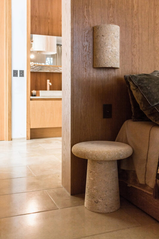 Stone stool beside a bed in a modern room with wooden walls and a visible bathroom in the background.