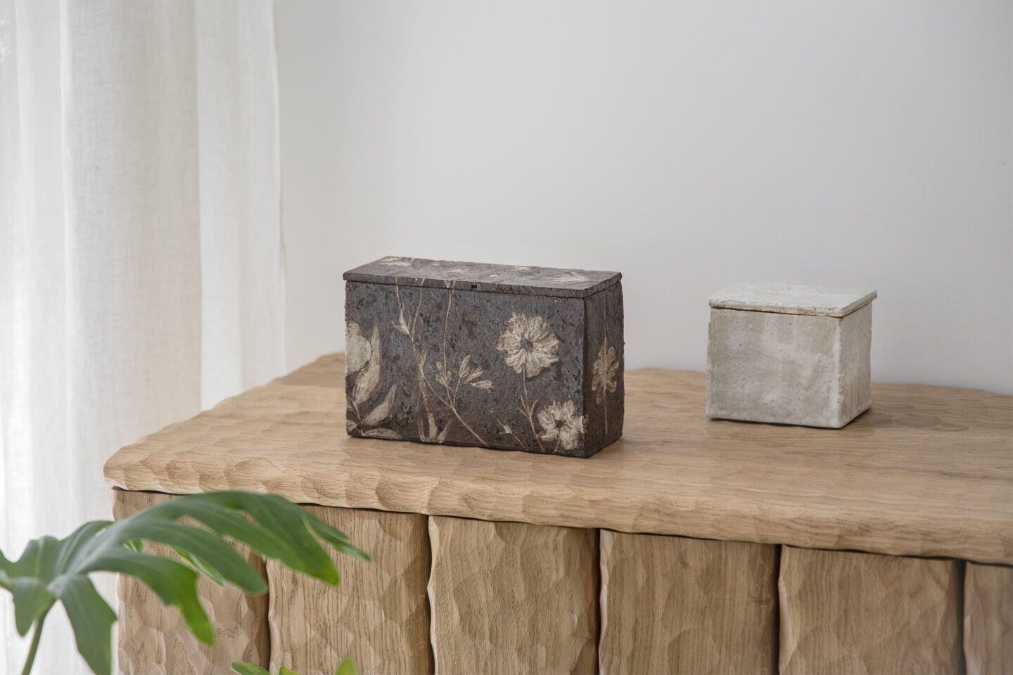 Decorative boxes with floral designs on a wooden surface, alongside a smaller plain box, with a plant leaf in the foreground.
