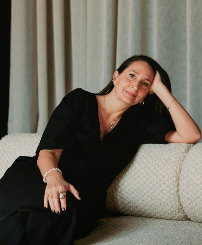Woman in a black outfit resting her head on her hand while sitting on a textured sofa, with soft drapes in the background.