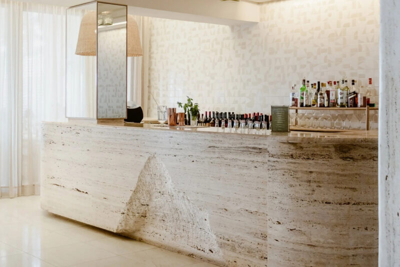 Modern bar with a textured stone counter, glass shelves displaying various bottles, and a light, airy atmosphere.
