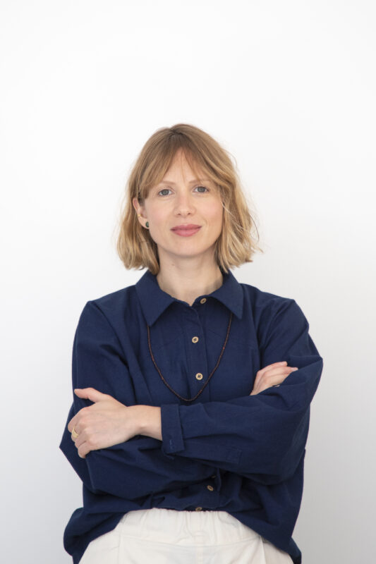 Woman with shoulder-length blonde hair, wearing a navy blue shirt, standing with arms crossed against a white background.