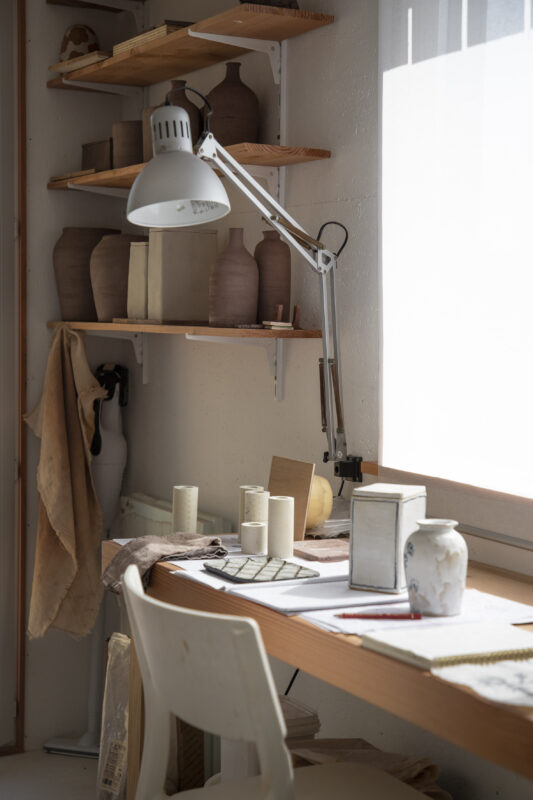 A well-lit workspace featuring a wooden table with pottery, tools, and a lamp, surrounded by shelves of ceramic pieces.