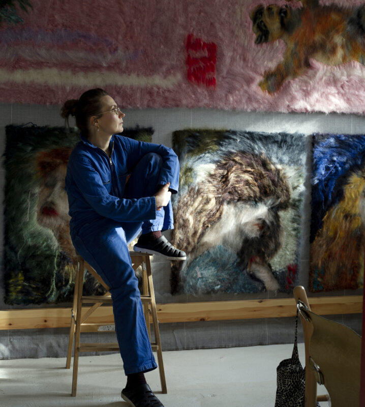 Artist Hanna-Kaisa Korolainen sits on a stool in a studio, surrounded by colorful, textured animal artworks.