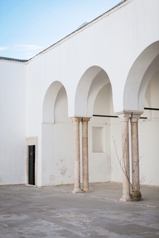 White-walled courtyard featuring arched columns and a potted plant, with a clear blue sky above.