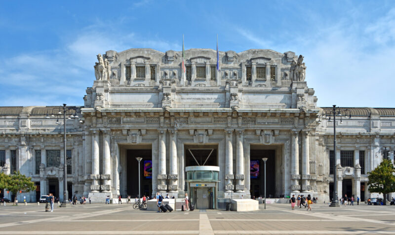 Grand façade of Milan Central Station with ornate architecture, flags, and people in the plaza under a clear blue sky.