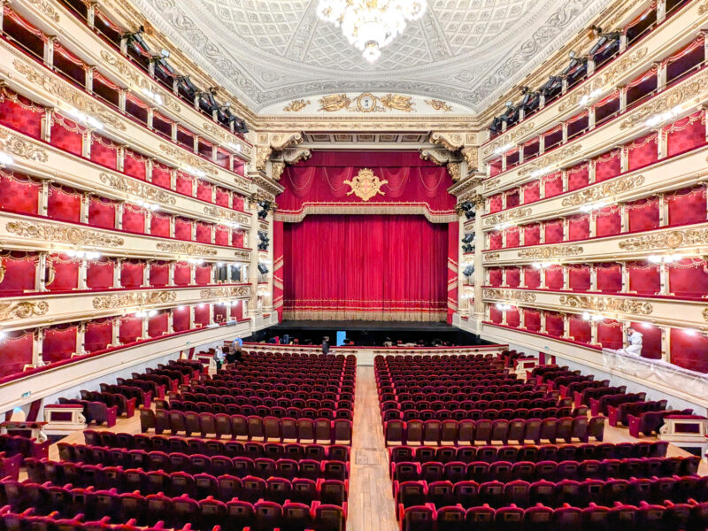 Interior view of Teatro alla Scala, showcasing red velvet seating and an ornate stage with a red curtain.
