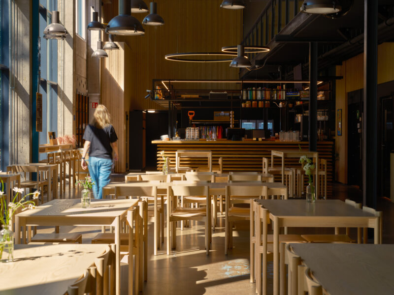 Brightly lit restaurant interior with wooden tables, a bar area, and a person walking towards the back.