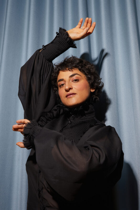 Portrait of a woman with short curly hair, wearing a black blouse, posing against a blue curtain backdrop.