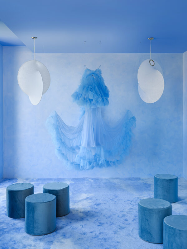 A light blue room featuring a tulle dress hanging on the wall, surrounded by circular stools and soft blue carpet.
