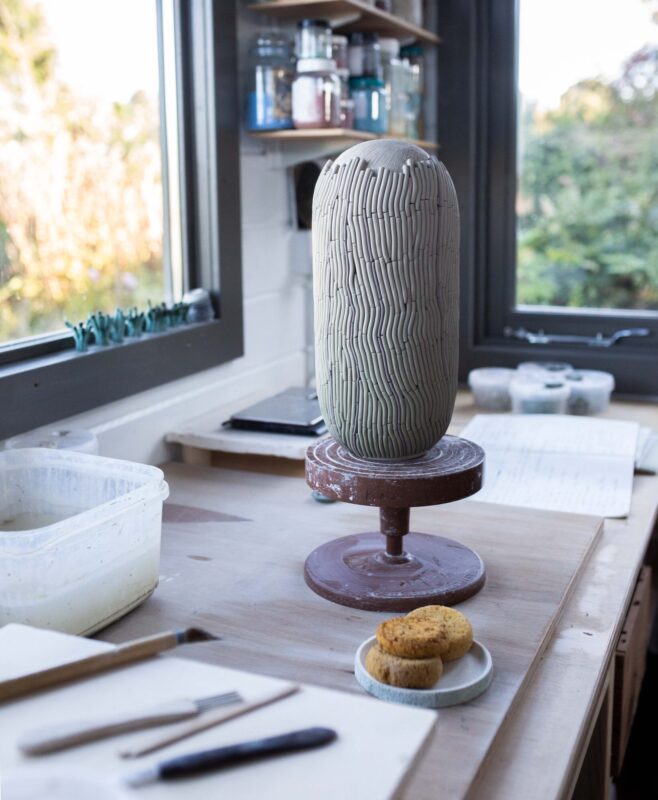 Ceramic vase with linear patterns on a pottery wheel, surrounded by tools and a plate of cookies in a studio setting.