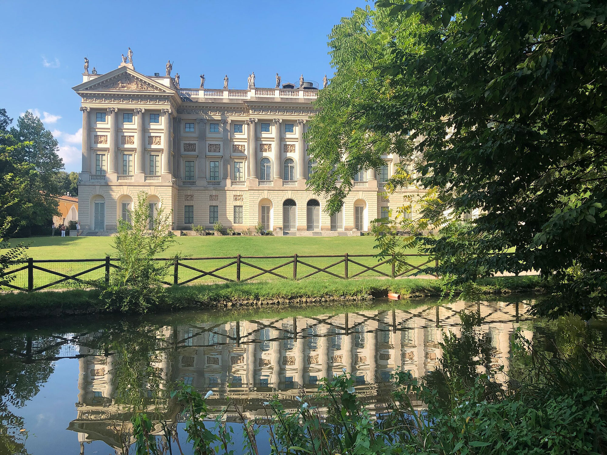 Villa Reale reflected in a calm waterway, surrounded by greenery and a wooden fence under a clear blue sky.