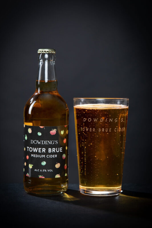 Bottle of Dowding's Tower Brue medium cider next to a glass filled with the cider, against a dark background.