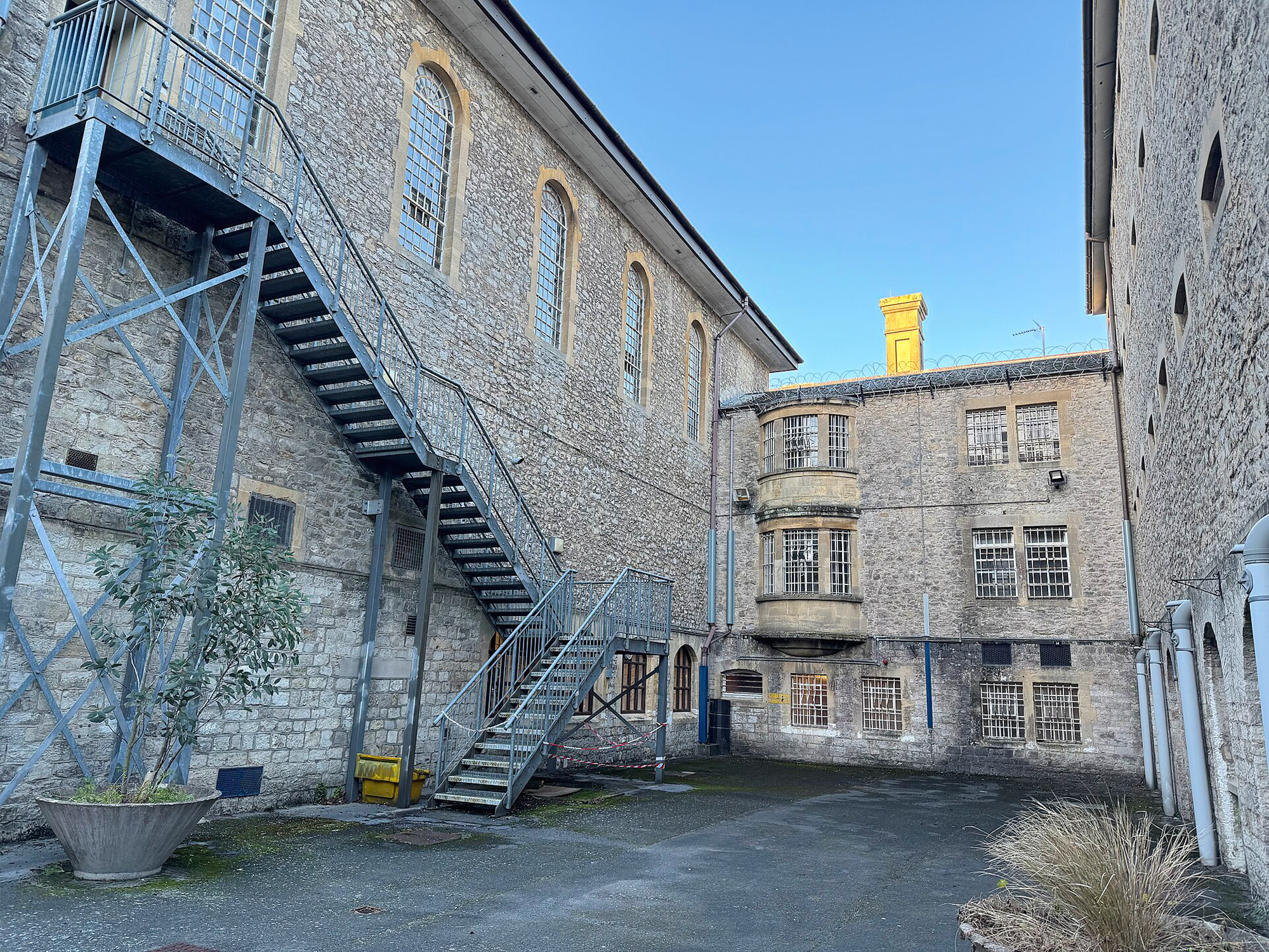 Courtyard of Shepton Mallet Prison featuring stone walls, a metal staircase, and barred windows.