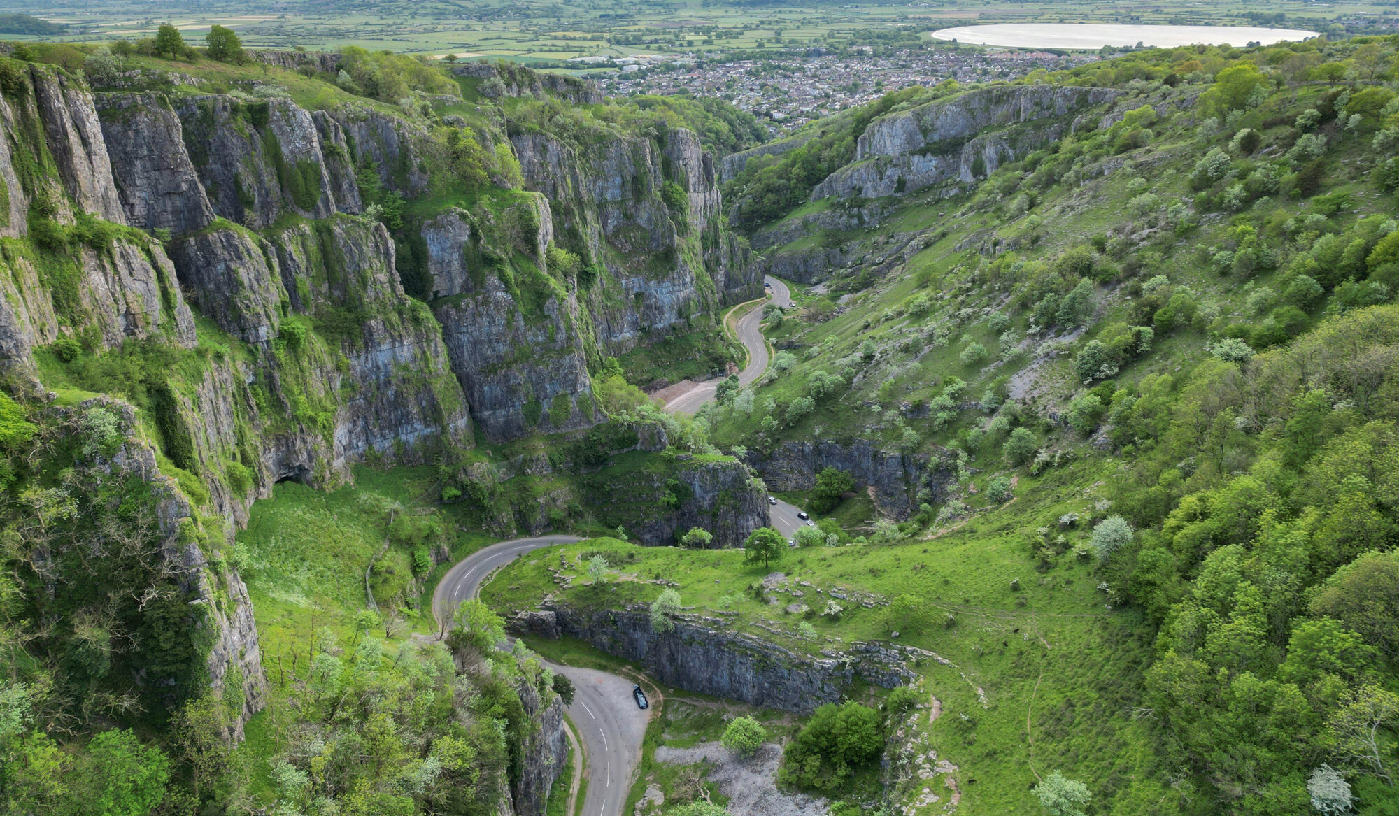 Lush green hills and rocky cliffs surround a winding road, with a distant view of a lake and cloudy sky.