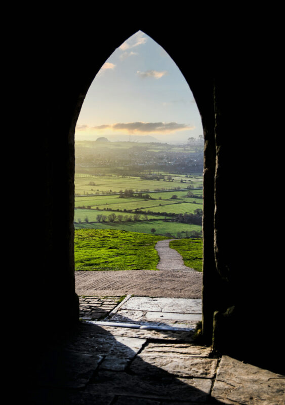 View through an arched doorway framing a lush green landscape and a winding path under a sunset sky.