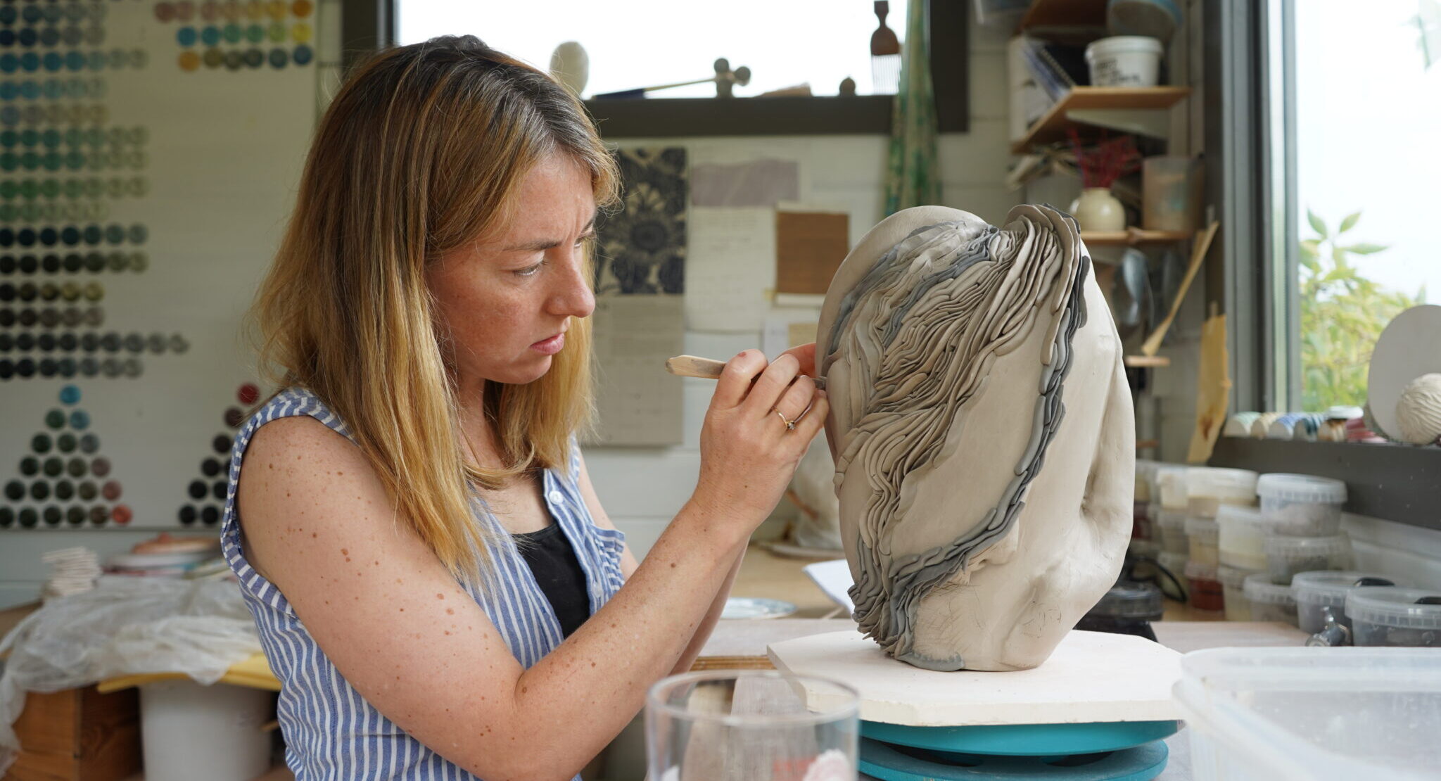 Artist sculpting a clay piece in a studio, surrounded by colorful materials and tools.