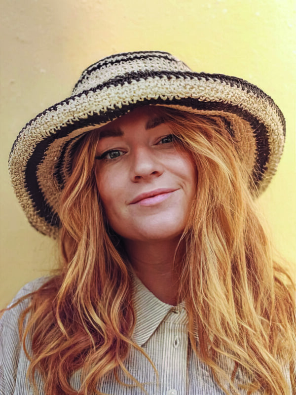 Woman with long, wavy red hair wearing a striped straw hat and a button-up shirt, smiling against a yellow background.