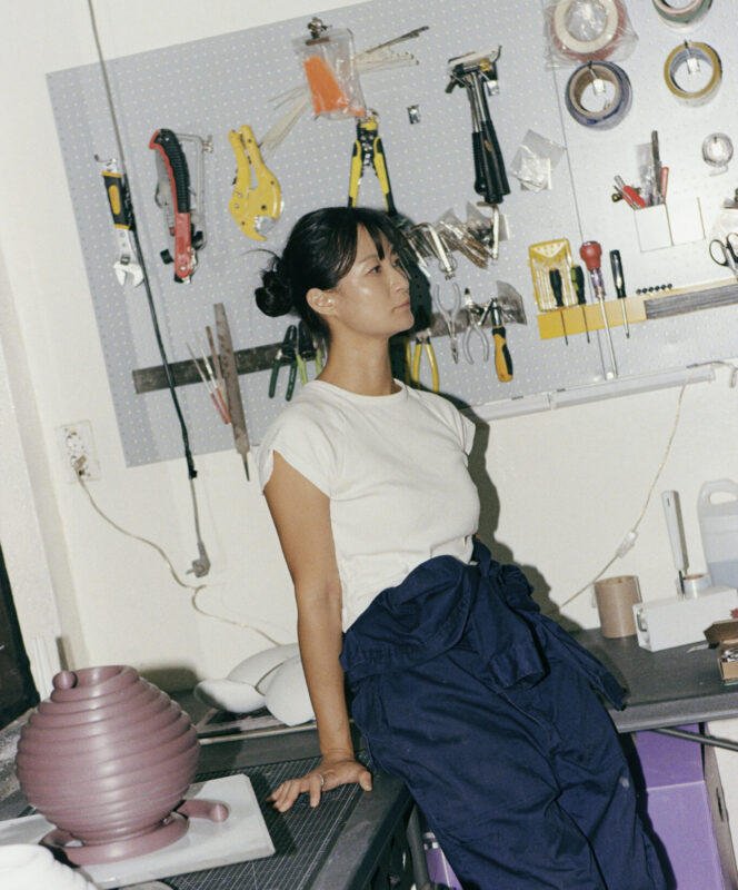 A woman in a white shirt and blue pants leans against a workbench with tools and materials in a creative office space.