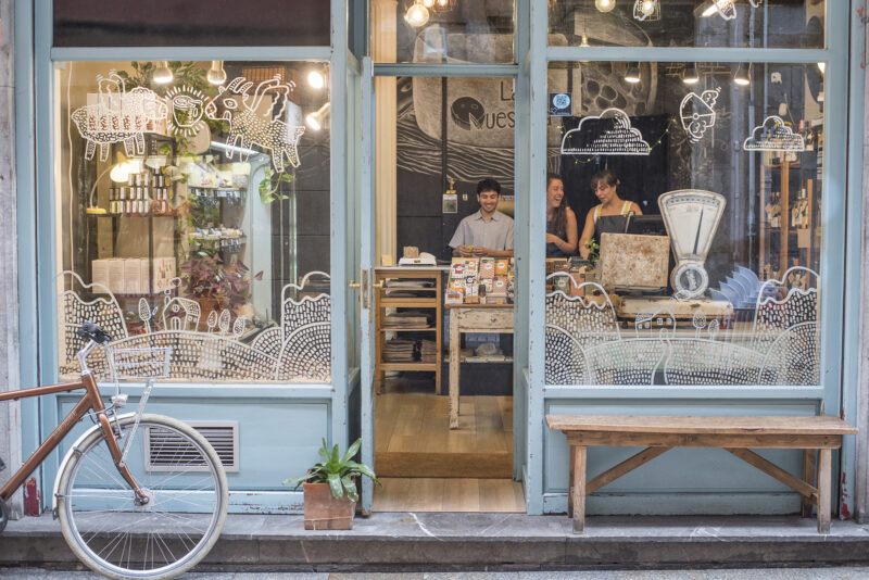 A quaint cheese shop with a bicycle outside, featuring large windows adorned with playful illustrations and two staff memb...