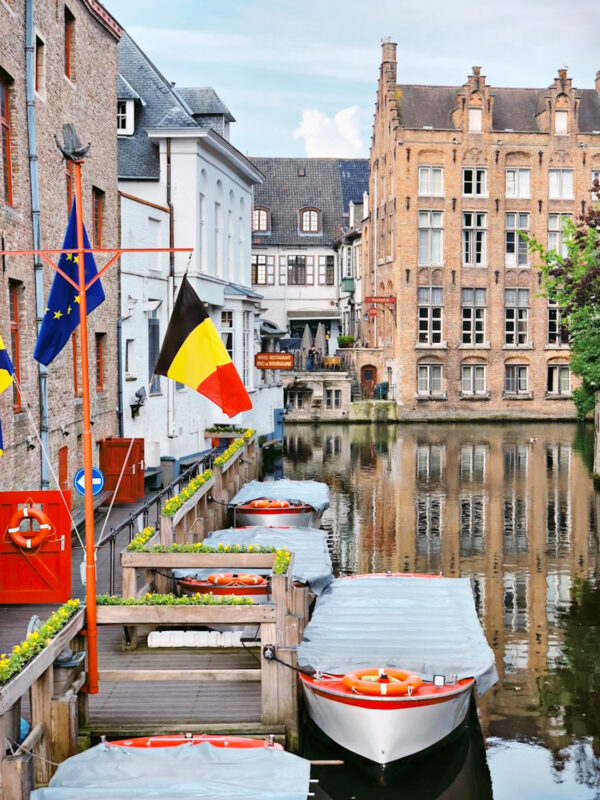 Canal view in Bruges with boats, Belgian flags, and historic buildings reflecting in the water.