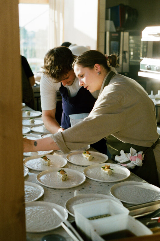Two chefs carefully plating dishes in a bright kitchen, focusing on presentation with white plates.