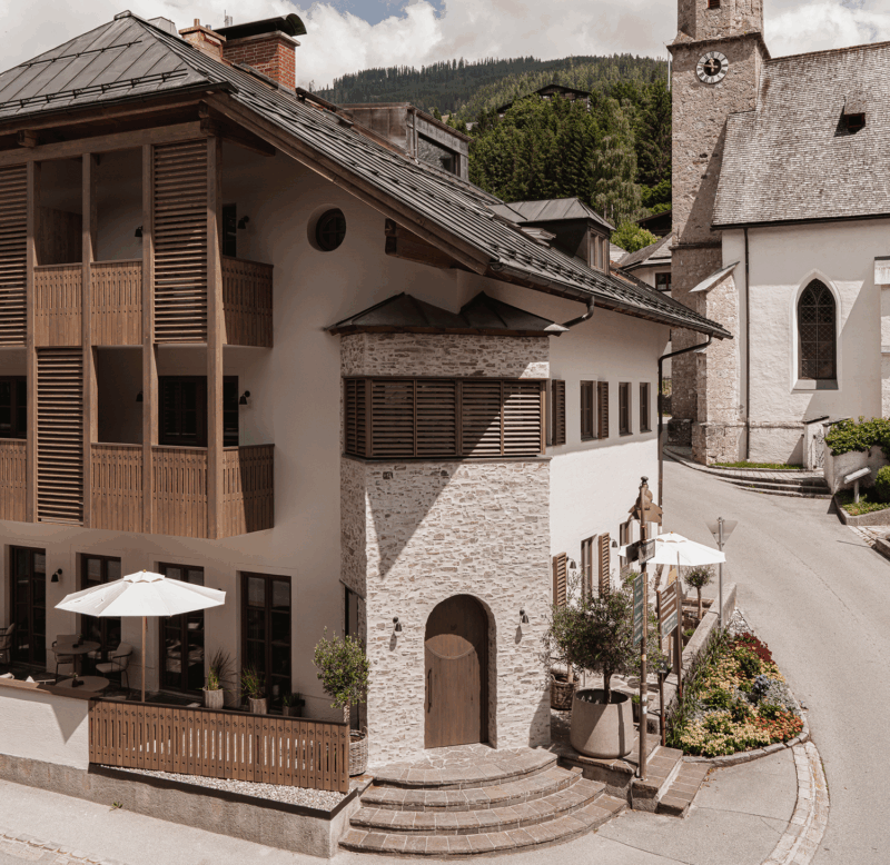 Charming building exterior with wooden balconies, stone accents, and umbrellas, set in a scenic village with mountains.