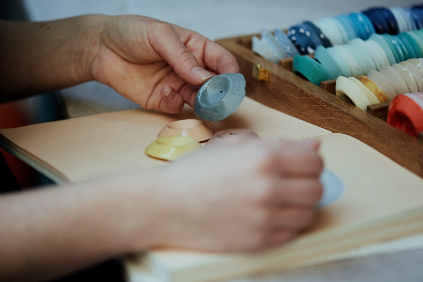 Hands holding colored ceramic pieces above an open notebook, with a wooden box of various colored samples in the background.