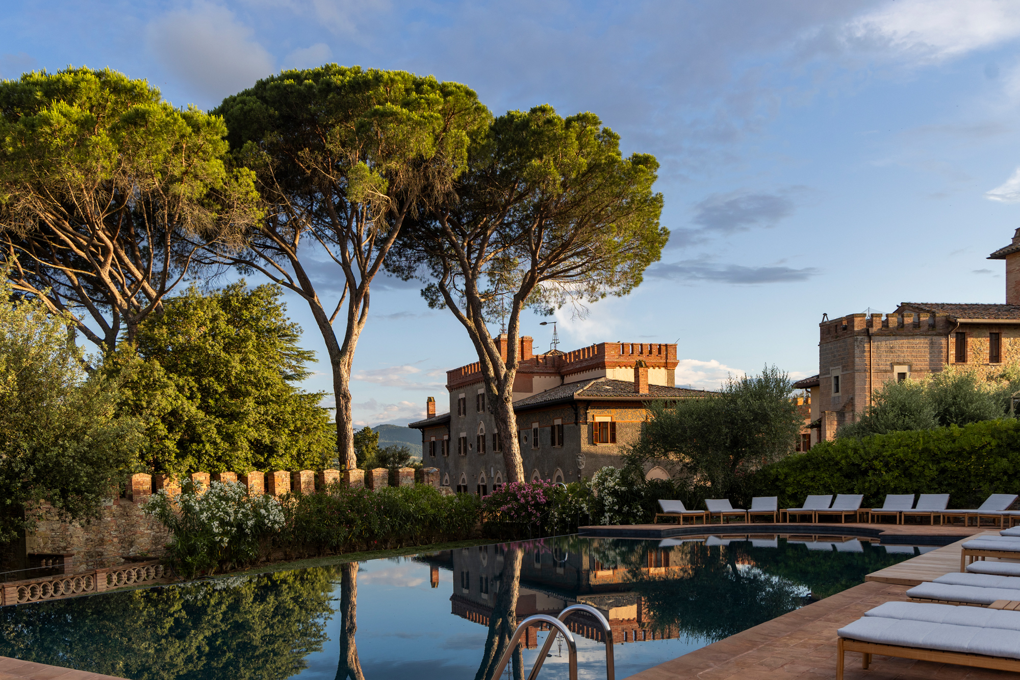 Serene swimming pool at Borgo Dei Conti Resort, surrounded by lush trees and historic architecture, reflecting the sky.