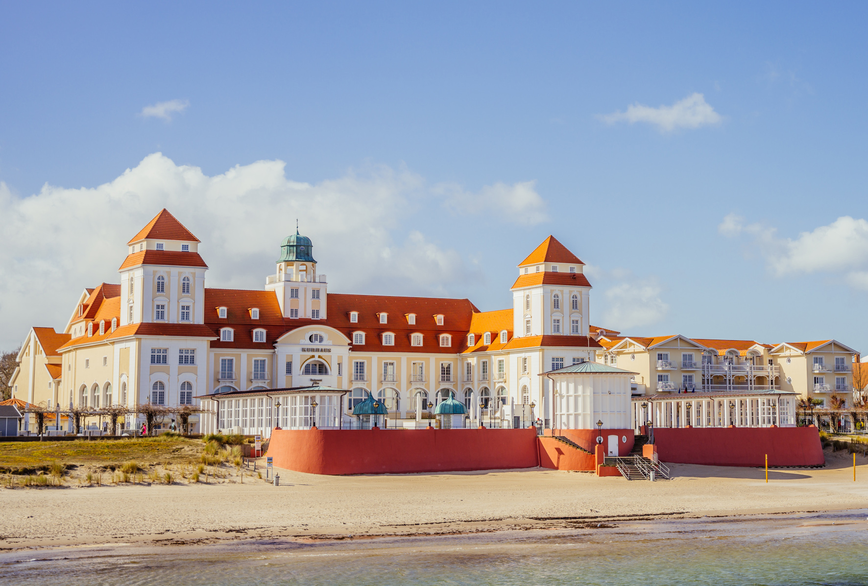 Kurhaus Binz, a large seaside building with red roofs, situated on the beach under a blue sky.