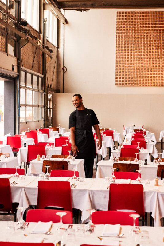 A smiling man in a black apron stands in a spacious restaurant with red chairs and neatly set tables.
