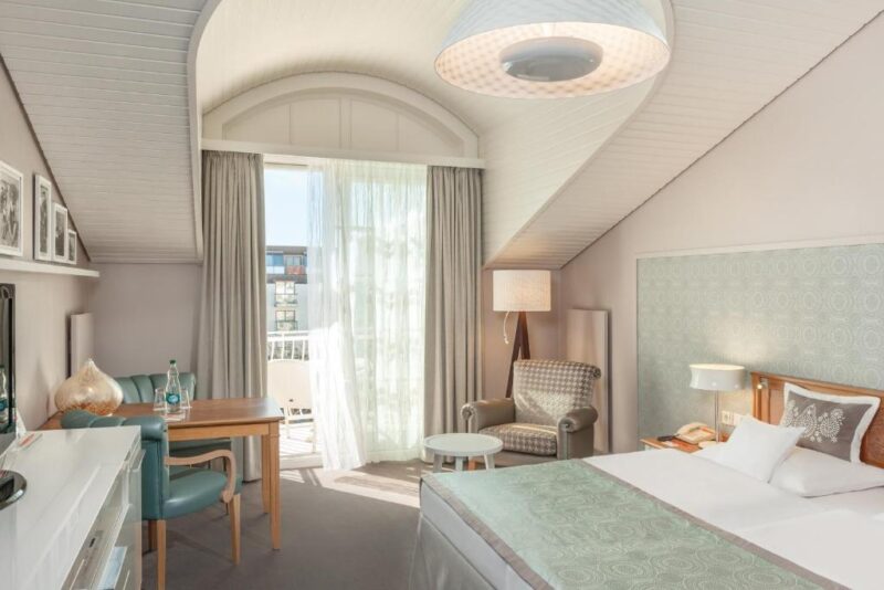 Bright hotel room with sloped ceiling, featuring a bed, seating area, and balcony access, uploaded to Kurhaus Binz.
