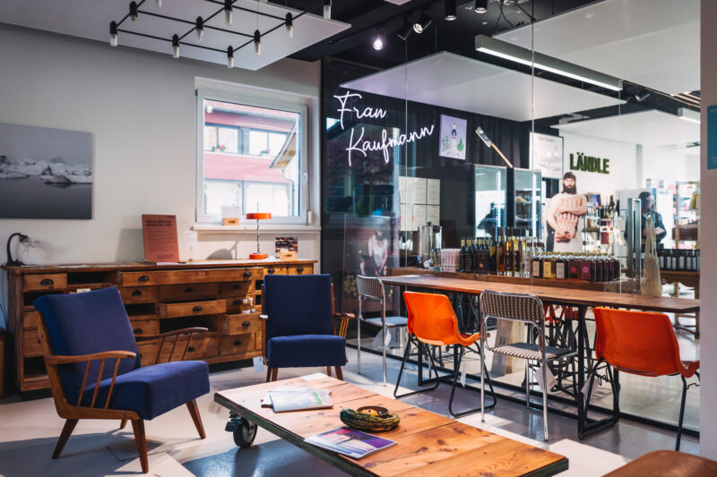 Modern café interior with wooden furniture, blue chairs, and orange accents; shelves filled with bottles in the background.