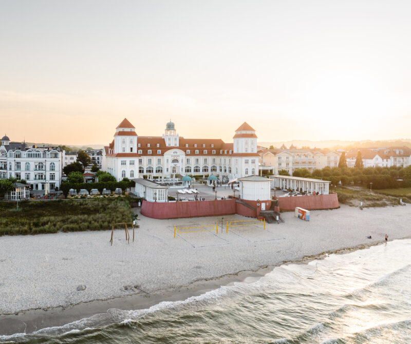Kurhaus Binz hotel with red-roofed towers, beach volleyball court, and sunset over the sea.