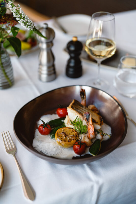 Gourmet dish featuring grilled fish, cherry tomatoes, and lemon, served in a dark bowl with wine and condiments nearby.
