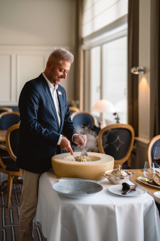 A man in a suit serves pasta from a large cheese wheel in an elegant restaurant setting.
