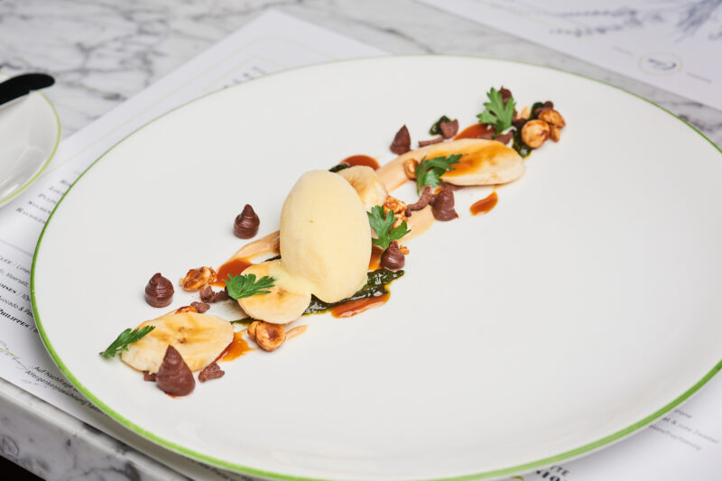 Dessert plate featuring banana slices, caramel sauce, chocolate dots, popcorn, and a scoop of banana sorbet, garnished wit...