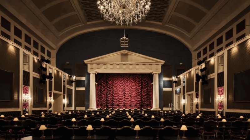 Interior of Kurhaus Binz, featuring a grand stage with red curtains and elegant chandeliers above rows of tables.