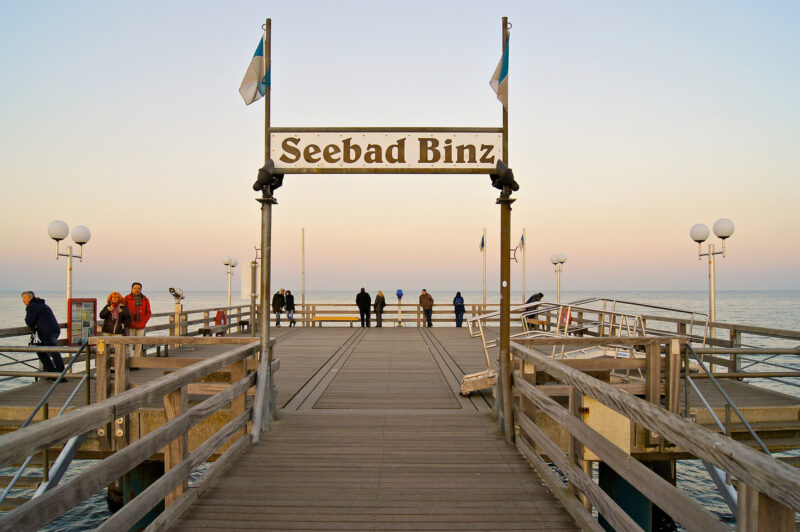 Wooden pier at Seebad Binz, with people walking and flags flying, against a pastel sunset sky.