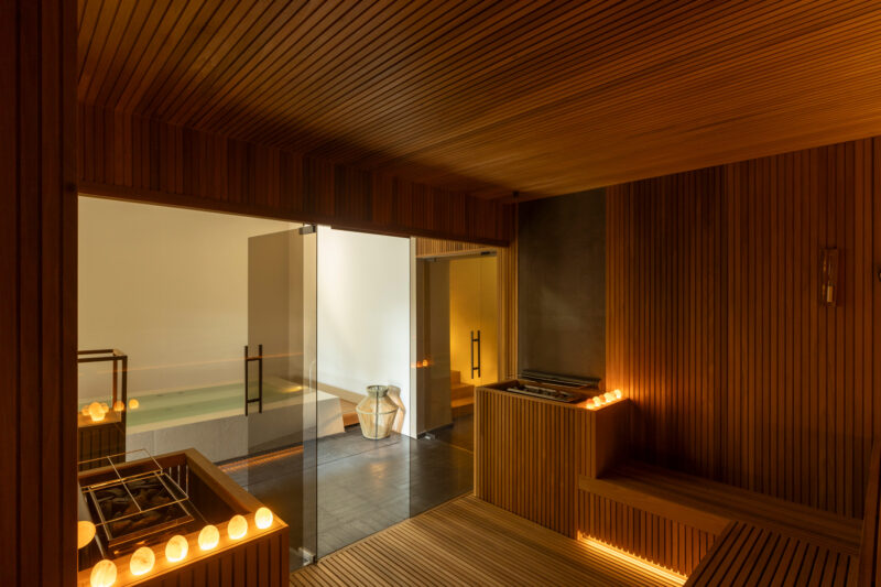 Finnish sauna interior with wooden paneling, dim lighting, and a glass door leading to a relaxation area.