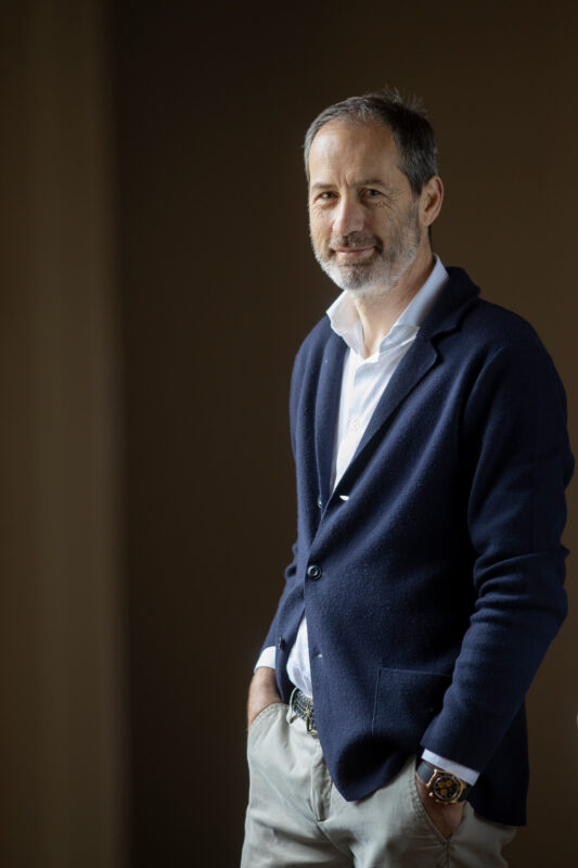 Man in a navy blazer and white shirt, smiling while standing with hands in pockets, against a neutral background.