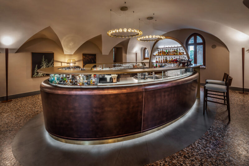 Elegant bar area at Borgo Dei Conti Resort, featuring a curved counter, shelves of liquor, and ambient lighting.
