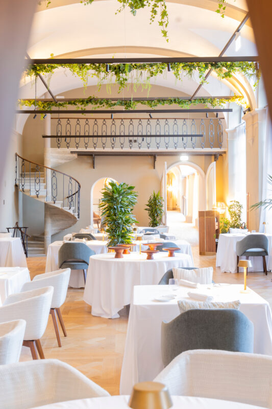 Elegant dining area at Borgo Dei Conti, featuring white tables, greenery, and a staircase in a bright, airy space.