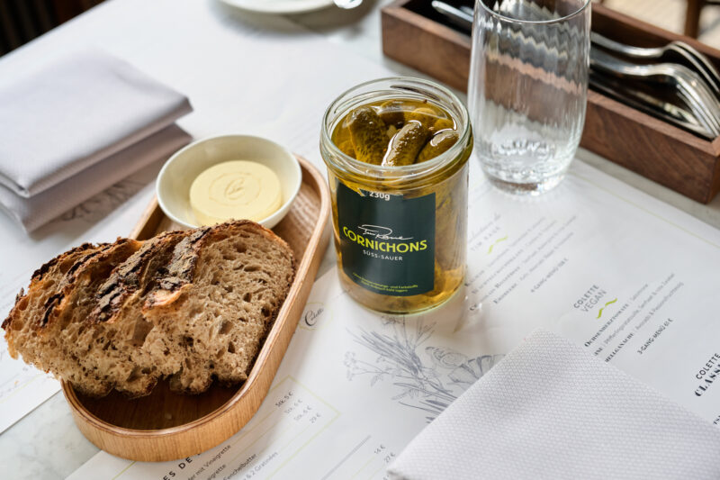 Sliced bread on a wooden plate with a jar of cornichons and butter on a table setting at Brasserie Colette.