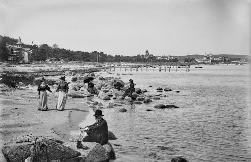 Historic black and white photo of people on a beach in Binz, Rügen, with rocky shoreline and distant buildings.
